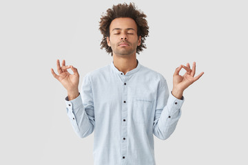 Attractive mixed race young male pracitices yoga, feels relaxed and calm, shows mudra sign with both hands, closes eyes as tries to concentrate on something, poses alone against white background