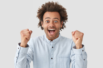 Energetic curly dark skinned young male has joyful expression, clenches fists with happiness, celebrates successful day, wears elegant white shirt, stands indoor. People, body language concept