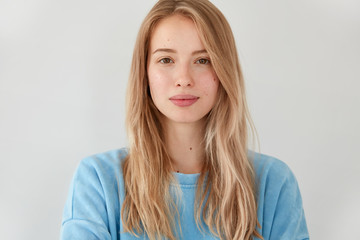 Close up shot of pleasant looking serious blonde Caucasian female with long hair, wears blue casual...