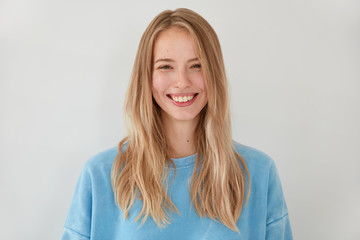 Pretty smiling blonde female with appealing tender look, has joyful expression, clean skin, being in high spirit after recieving compliment from handsome guy, stands against white background.