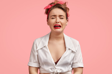 Depressed European female in low spirit, cries desperately, has red lips closes eyes, has grief, wears white top and headband, stands against pink wall. Lonely stressful woman in retro style