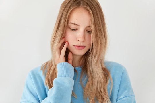 Cropped shot of beautiful young European female with staright hair, looks thoughtfully down, has sad pensive facial expression, dressed in blue oversized sweater, isolated over white background