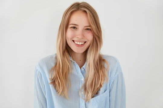 Cheerful blonde young female with attractive look, broad shining smile, dressed in fashionable shirt, stands against white background, being in high spirit after stroll during sunny spring day