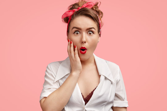 Emotive pinup girl with stupefied expression, being amazed by sudden news, opens mouth widely, can`t believe in fault, isolated over pink background. Surprised young woman in retro style clothes
