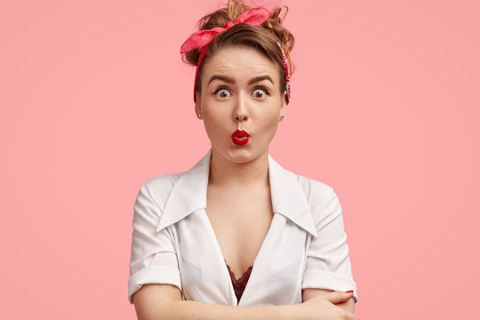 Stunned young female with surprised expression, keeps red lips round, hands crossed, shocked as notices something, dressed casually, isolated over pink background. People, facial expressions concept
