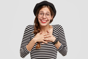 Attractive positive female dressed in French style, keeps both palms on heart, being touched by recieving compliment, has broad smile, red lipstick, stands against white studio wall. Woman from France