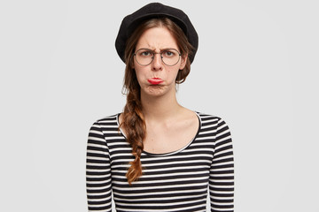 People and insult concept. Beautiful young Parisian female being abused by negative news, purses lower lip, wears casual clothes in French style, isolated over white background, has problems in life