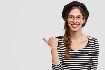 Indoor shot of friendly Parisian female with broad smile, points on left aside at blank copy space, dressed in French style, invites you to visit cosy cafe in France, isolated over white background