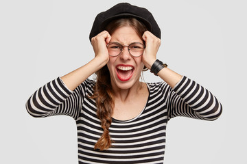 Depressed beautiful young woman dressed in French style, keeps hands on head, looks desperately at camera, feels worried and stressed, has many problems, stands against white studio blank wall.