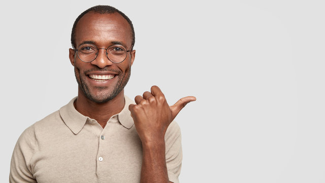 Indoor shot of pleasant looking dark skinned male with cheerful expression, points aside with thumb, has broad smile, advertises something, isolated on white background. Handsome African American man