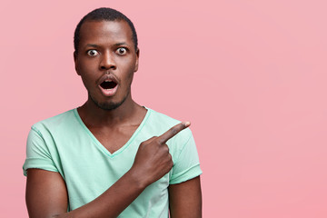 Emotional dark skinned male with astonished expression, has shocked look, indicates aside with fore...