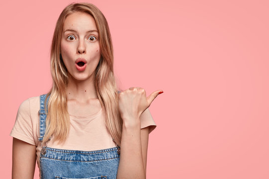 Surprised beautiful young female with shocked expression, points aside with thumb, keeps jaw dropped, dressed in denim overalls, shows blank space for your advertisement or promotional text.