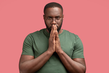 Studio shot of serious black male keeps hands in praying gesture, worships for something, hopes everything will be good, has stong body, wears t shirt, isolated over pink background. God, help me!