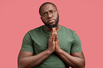 Photo of serious African American male with pleading expression, keeps palms pressed together, asks for something his lover, hopes to recieve positive answer dressed in casual outfit, isolated on pink