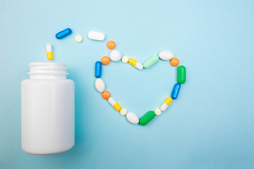 Multi-colored tablets and heart-shaped capsules, white bottle for tablets, pharmaceutical medicine pills on blue background, an analgesic against diseases