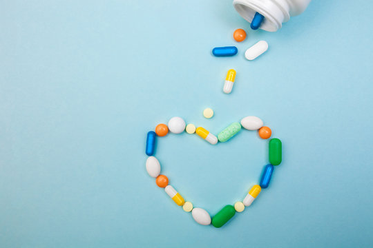 Multi-colored tablets and heart-shaped capsules, white bottle for tablets, pharmaceutical medicine pills on blue background, an analgesic against diseases