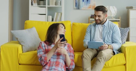 Caucasian pretty woman sitting on the floor at home with a smartphone in hands and her boyfriend sitting on the couch with a tablet computer.