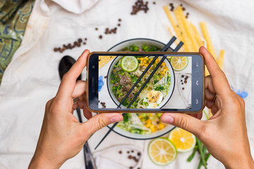 Smartphone food photography. Woman hands take phone photo. Ramen noodle soup with egg, corn and meat in bowl. Asian Thai traditional authentic food with vegetables served in cafe or restaurant