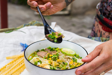 Fototapeta na wymiar Woman hand holds and eat ramen noodle soup with egg, corn and meat in bowl. Asian Thai traditional authentic food with vegetables served in cafe or restaurant