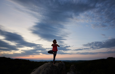 Obraz premium Full-length picture of young slim fit woman doing yoga exercises standing barefooted on one leg on big mountain rock against beautiful evening sky. Fitness and active lifestyle concept. Vrikshasana