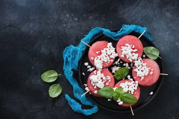 Obraz na płótnie Canvas Above view of a metal tray with round watermelon slices on skewers and cottage cheese, horizontal shot over dark scratched metal background with space