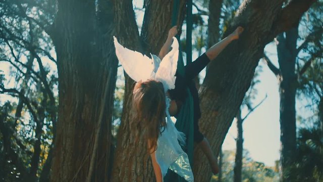 A woman in a fairy costume performing aerial silks hanging from a tree in slow motion.