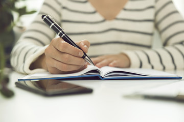 Young business woman sitting at table and writing in notebook. On table is smartphone, and tablet....