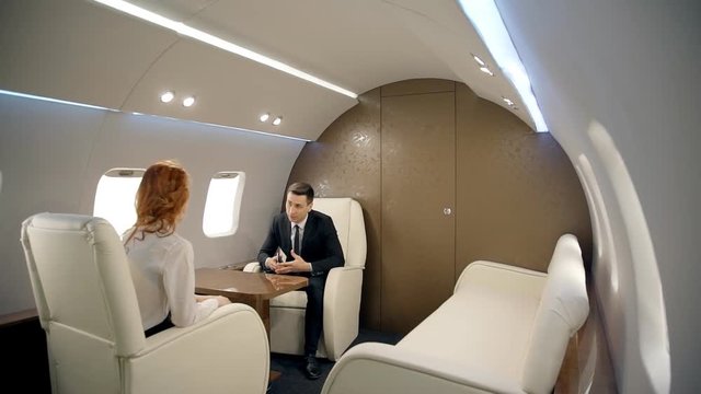 Young businesspeople are discussing modern business, sitting in private plane, stylish businessman in black suit is talking with businesswoman during flight in luxury interior. Concept: conversation