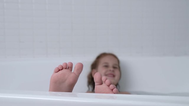 Small girl is in bathtub. Camera is focused in the child feet located on the foreground. Washing her face with the hands on the background.