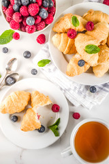 Sweet summer dessert, homemade baked mini croissants with berry jam, served with tea, fresh raspberries, blueberries and mint. On a white marble table, copy space top view