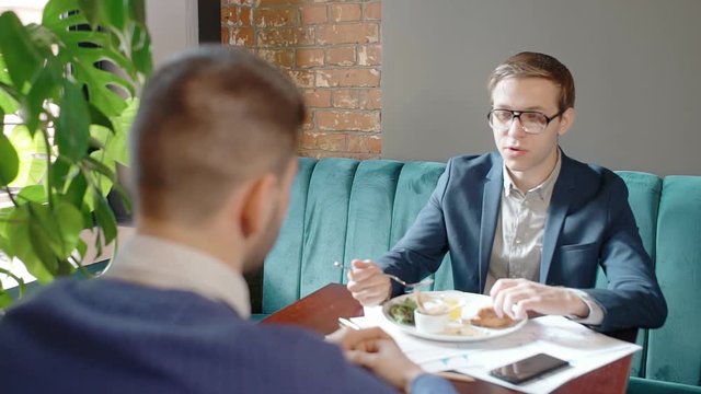 Young businessmen are talking during business lunch, sitting at table in restaurant, two successful men discussing startup project at desk, enjoying delicious meal in modern interior. Concept