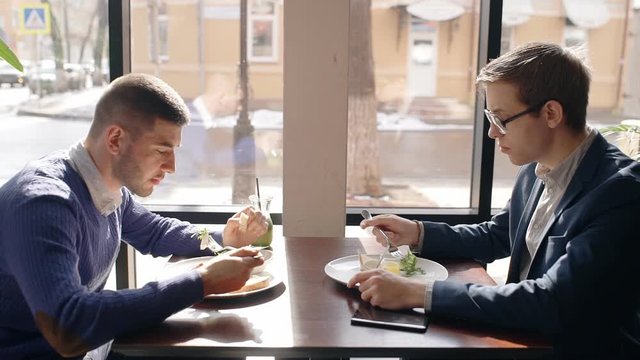 Two Young Men Are Eating And Talking, Sitting At Table In Cafe, Handsome Guys Is In Dialogue During Business Lunch At Desk In Bright Room By Window With Urban View. Concept: Friendship, Business