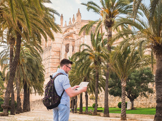 Attractive man with sunglasses and a backpack, looking at the map with sights on the background of palm trees and historical buildings. Spain, Palma de Mallorca © Svetlana