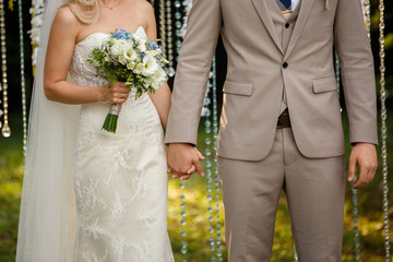 Close-up view of stylish groom and bride with bouquet in beautiful dress. Unrecognizable newlyweds holding each other hands. Outdoors.