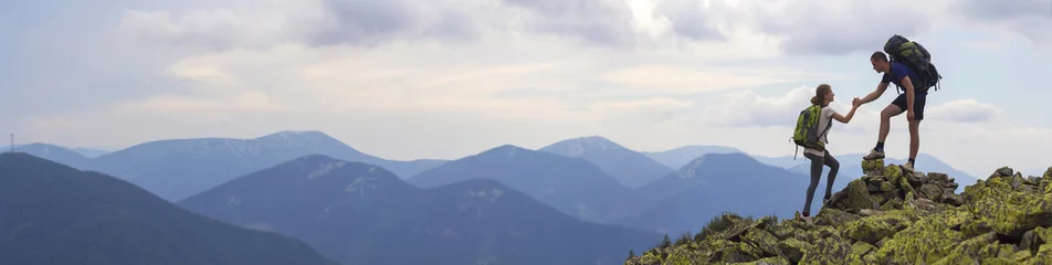 Deurstickers Jonge toeristen met rugzakken, atletische jongen helpt slank meisje om de rotsachtige bergtop te beklimmen tegen een heldere zomerhemel en bergketen. Toerisme, reizen en gezonde levensstijl concept. © bilanol