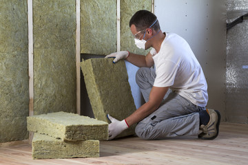 Worker in protective goggles and respirator insulating rock wool insulation in wooden frame for future house walls for cold barrier. Comfortable warm home, economy, construction and renovation concept