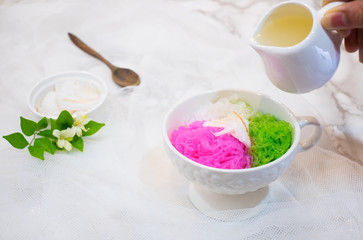 Women hand pouring coconut milk sauce on Salim (colorful sweet noodles) for summer time on white marble background with wooden spoon. Special taste of Thai dessert.
