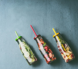 Colorful infused water in bottles with fruits berries, cucumber, herbs and drink straws on gray background, top view. Tasty summer clean beverages for  healthy lifestyle and fitness