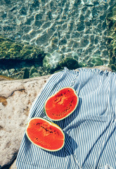 Two watermelon halfs are on striped carpet. Sea lagune with clear water, rocky bank