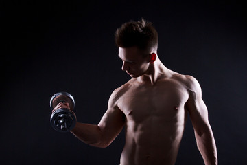 Fototapeta na wymiar Athletic young man with a dumbbell on a black background. Naked torso, muscular body. Strong chest and shoulder muscles. Studio shot, low key. Bodybuilding concept