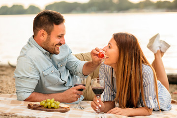 Happy young couple having a picnic at the beach. Lying on the blanket and drinking wine. Boyfriend giving cherry tomato to his girlfriend