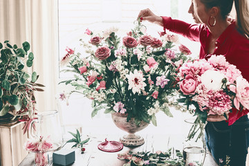 Florist women make beautiful big festive event classical bouquet with roses and other flowers in urn vase on table at window, lifestyle
