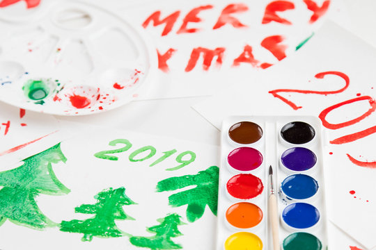 Children's drawings set by a watercolor on white paper of a congratulation by New year and Christmas. figure 2019 and three fir-trees largely green in color. A palette and a set of paints on a table. 