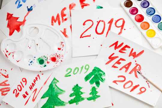 Children's drawings set by a watercolor on white paper of a congratulation by New year and Christmas. figure 2019 largely green and red in color. A palette and a set of paints on a table. Horizontal s