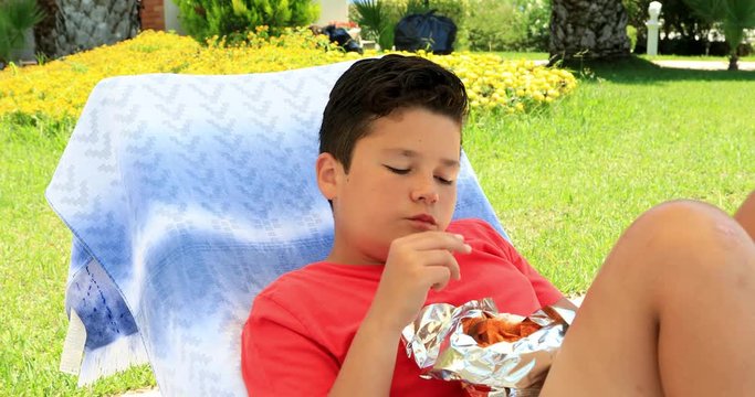 Portrait of a cute preteen schoolboy laying on a sunbed and eating potato chips