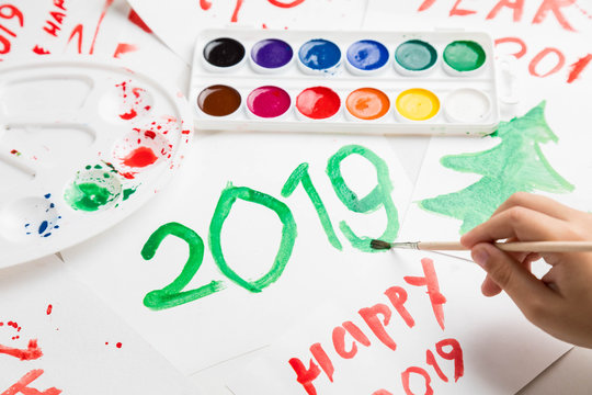Children's drawings set by a watercolor on white paper of a congratulation by New year and Christmas. Figure 2019 largely green paint. A palette and a set of paints on a table. Horizontal shot