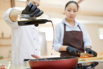 Mid section of two professional cooks working in restaurant kitchen poring soy sauce into frying...