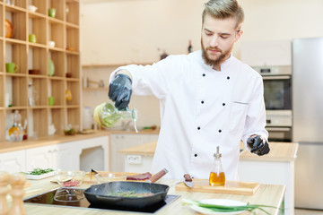Portrait of handsome  professional chef working in modern restaurant kitchen standing at wooden table and adding olive oil to delicious dishes, copy space