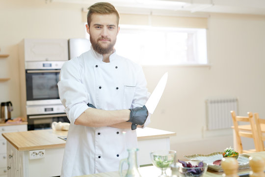 Portrait of handsome bearded chef posing in kitchen standing with arm crossed at wooden table and looking at camera, copy space