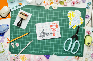 Multi-colored paper, homemade postcard and scrapbooking tools and materials on green mat for cutting, top view, no hands